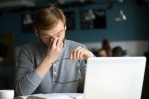 Man rubbing his eyes while using a laptop, a common sign of blurry vision or eye strain.