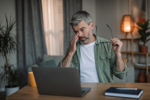 Man rubbing his irritated eye while working on a laptop, illustrating dry eye symptoms associated with screen time.